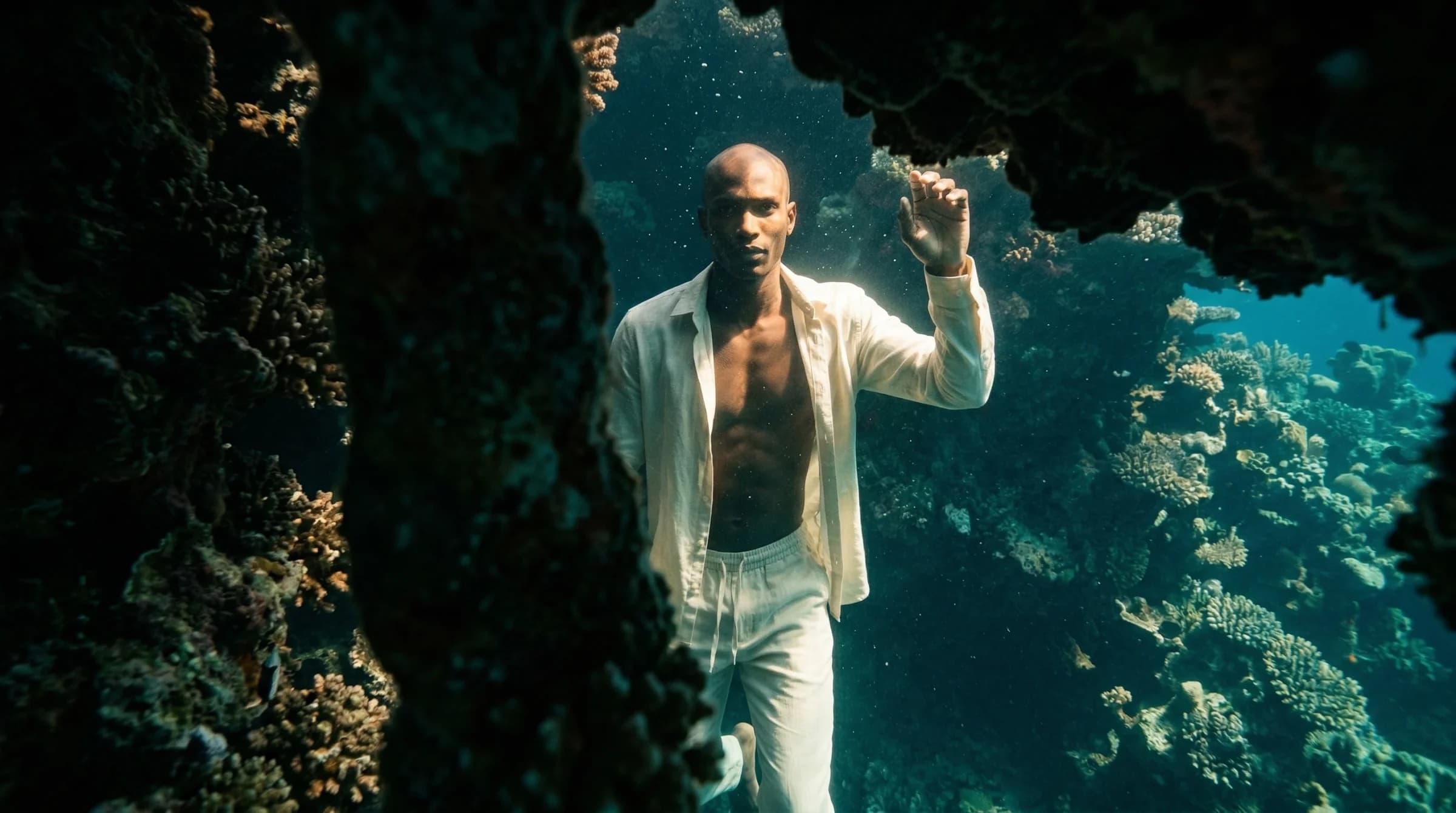 A figure in cream linen emerging from reef shadow into a single shaft of sunlight underwater